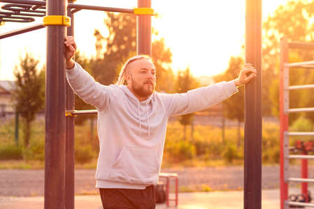 A man with a beard and an interesting hairstyle is leaning on a horizontal bar in the morning on the sports ground.の写真素材
