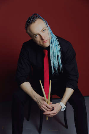 A young attractive man with an interesting hairstyle in pigtails on an undercut with a red tie on a red background in and drumsticks in his hands.の写真素材