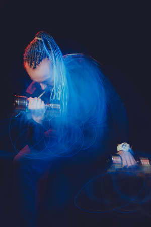 A handsome man with an interesting hairstyle from braids on a black background shakes his biceps...