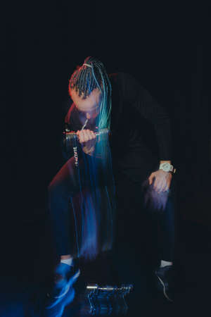 A handsome man with an interesting hairstyle from braids on a black background shakes his biceps with dumbbells 5 kilograms, shooting with mixed light.の写真素材