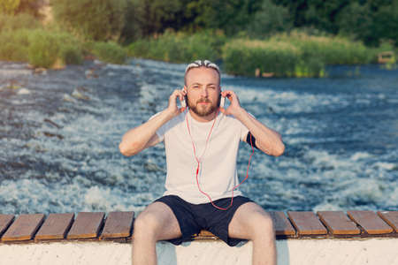 A man with a beard and a braid haircut listens to music with headphones on the river bankの写真素材