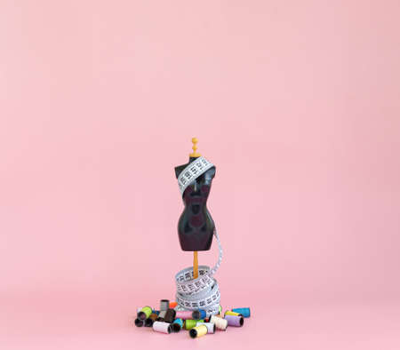 A scaled-down mannequin on a pink background with a tape for measuring and multicolored threads in spoolsの写真素材