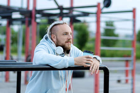 A man with a braid hairstyle in the early morning drinks coffee on the sports ground and listens to music on headphones.の写真素材