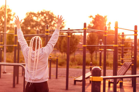 In the morning at sunrise, a man with an interesting braid hairstyle triumphantly raised his hands up after a workout. Stands with his back to the camera.の写真素材