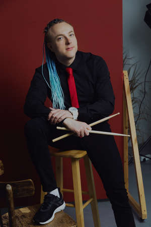 A young attractive man with an interesting hairstyle in pigtails on an undercut with a red tie on a red background in and drumsticks in his hands.の写真素材