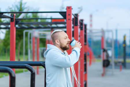 A man with a braid hairstyle in the early morning drinks coffee on the sports ground.
