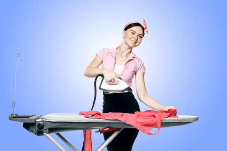 A beautiful girl dressed in pin-up style is ironing clothes isolated on a blue background.の写真素材