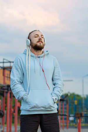 A man with a braid hairstyle in the early morning listens to music with headphones on the sports field against the background of the pink-blue sky.の写真素材