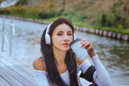 A woman in sportswear listens to music on headphones after a workout and drinks coffee on the river bank.の写真素材