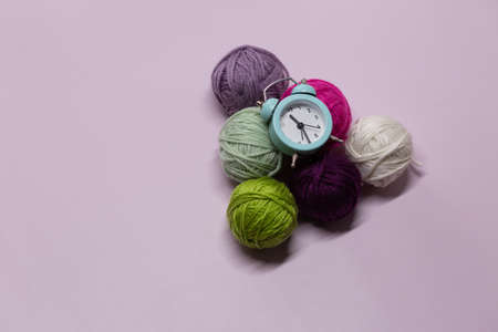 A small green alarm clock lies on woolen balls against a lilac background. Time to knit.