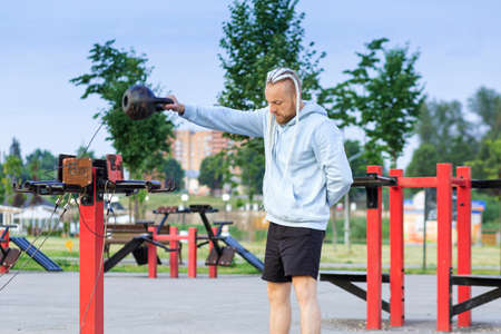 A man with an interesting braid hairstyle in a blue hoodie works out early in the morning on the street and makes swings with a kettlebell.の写真素材