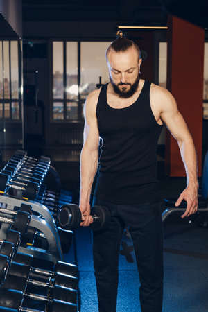A man of athletic build poses against the background of a rack with dumbbells in the gym.