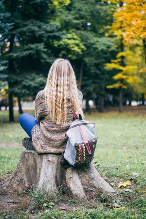 A girl sitting on a stump with a backpack. The view from the back