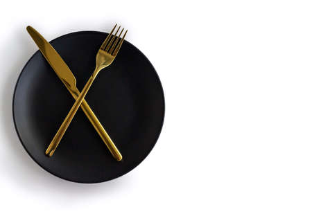Black empty ceramic plate with golden cutlery on a white background. Center composition. copyspace.