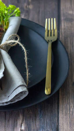 Table setting with black ceramic plate, golden fork, linen napkin on the wooden table. cropped...