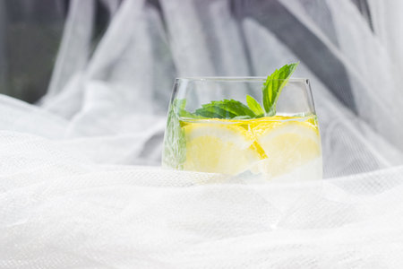 Close-up of homemade lemonade with lemon, mint and ice cubes in a glass on white wood table with white tulle fabricの写真素材