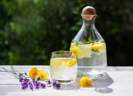 Homemade lemonade with lemon and ice cubes in a glass pitcher on the white wood table with yellow flowers and lavenderの写真素材