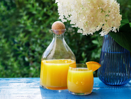 Fresh orange juice in pitcher and a glass garnished with a slice of ripe orange on blue wooden table with flowersの写真素材