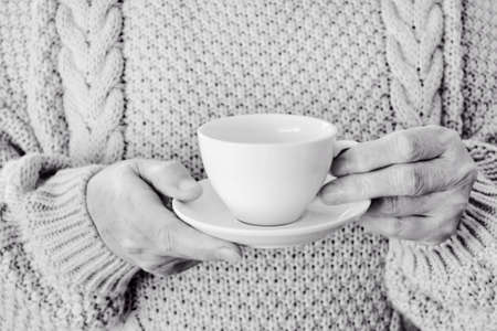 Elderly hands of a woman hold a white cup and saucer with tea at home close-up against the background of a knitted sweater.の写真素材