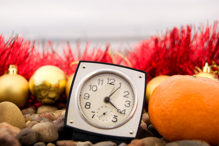 Christmas toys - golden balls, clock and tangerine on the beach, blurred sea and Christmas red tinsel in the background. New Year or Christmas card, invitation with copy space for text.の写真素材