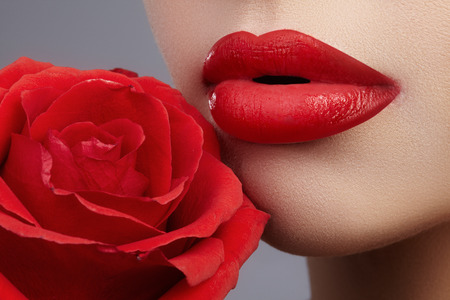 Close-up beautiful female lips with bright lipgloss makeup. Perfect clean skin, sexy red lip make-up. Beautiful visage portrait with red rose flower. Visage and cosmeticsの写真素材