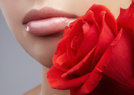 Close-up beautiful female lips with bright lipgloss makeup. Perfect clean skin, sexy pink lip make-up. Beautiful visage portrait with red rose flower. Visage and cosmeticsの写真素材
