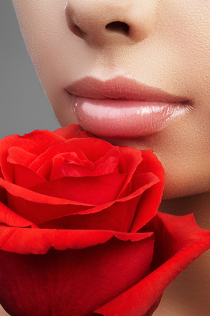 Close-up beautiful female lips with bright lipgloss makeup. Perfect clean skin, sexy pink lip make-up. Beautiful visage portrait with red rose flower. Visage and cosmeticsの写真素材