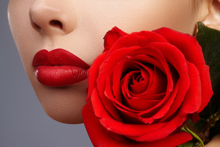 Close-up beautiful female lips with bright lipgloss makeup. Perfect clean skin, sexy red lip make-up. Beautiful visage portrait with red rose flower. Visage and cosmeticsの写真素材