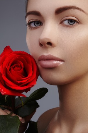 Close-up beautiful female lips with bright lipgloss makeup. Perfect clean skin, sexy red lip make-up. Beautiful visage portrait with red rose flower. Visage and cosmeticsの写真素材