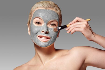 Beautiful Woman Applying White Facial Mask. Beauty Treatments. Close-up Portrait of Spa Girl Apply Clay Facial mask with Brush on grey backgroundの写真素材