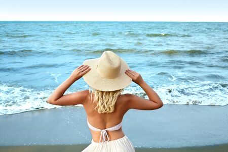 Blonde Woman in Fashion Summer Style Standing at Sea and Holding Hat. Luxury Lifestyle Rear View. Summer Vacation Concept with Ocean.の写真素材