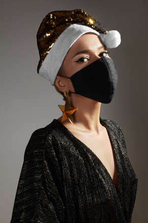 Woman with gold christmas hat, black medical mask. Fashion style with shiny clothes and accessories for xmas time. Protect yourself from Coronavirus Covid-19 even on holidaysの写真素材