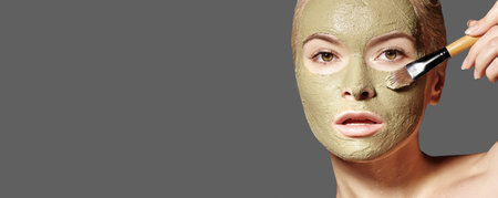 Beautiful Woman Applying Green Facial Mask. Beauty Treatments. Close-up Portrait of Spa Girl Apply Clay Facial mask on gray background.の写真素材