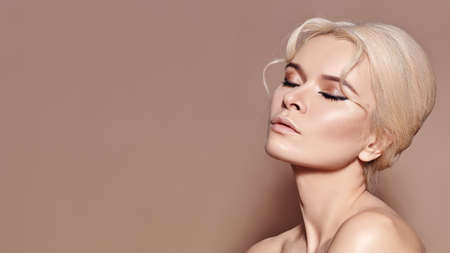 Beautiful sexy model with retro style make-up, clean skin, blond hair bun on beige background. Elegant woman model in femininity pose with fashion make-up, hairstyleの写真素材