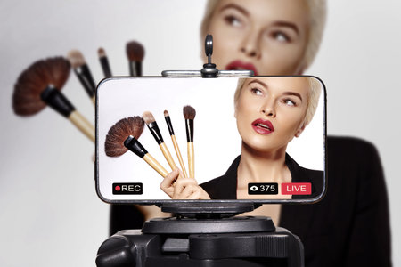 Confident young woman demonstrating how to apply makeup on herself during a live beauty tutorial. Female influencer using brushes and cosmetics while teaching makeup techniques for her online audienceの写真素材