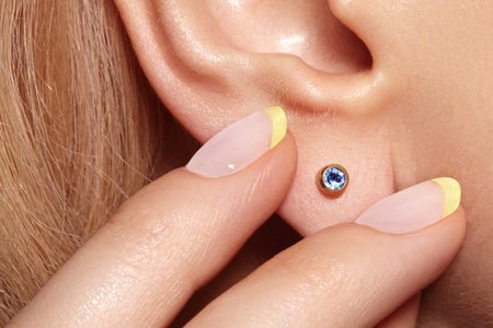 Close-up macro shot of female ear with gold stud earring featuring round blue gemstone. Elegant minimalist jewelry detail with polished metal and sparkling stoneの写真素材