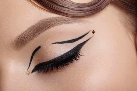 Close-up macro of a closed female eye with black winged eyeliner and tiny gold dot embellishment, soft lighting highlighting lash curve and eyelid decoration for editorial beauty useの写真素材