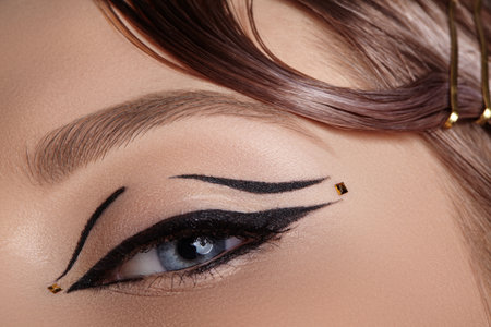 Close-up macro of a closed female eye with black winged eyeliner and tiny gold dot embellishment, soft lighting highlighting lash curve and eyelid decoration for editorial beauty useの写真素材