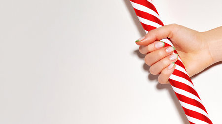 Beautiful woman palm with french manicure on nails holds red candy cane. Nails art design. Close-up of hands with trendy neon nails on striped print. Christmas in design for beauty and cosmeticの写真素材