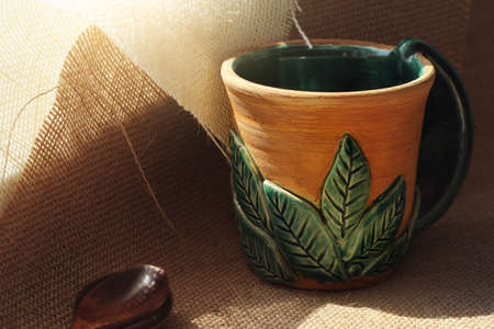 Hand made ceramic mug with leaves on brown burlap illuminated by the rays of the sun. Dark wooden spoons. The concept of pottery art and rustic style.の写真素材