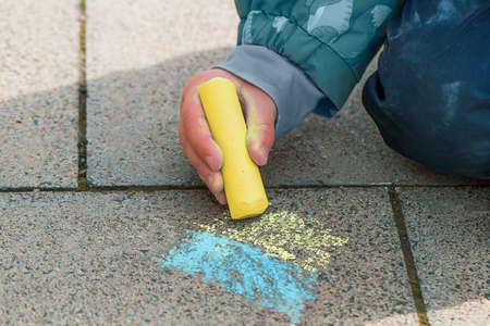 The child draws the Ukrainian flag on the pavement. A child's hand is holding a yellow chalk. Ukraine support banner.の写真素材