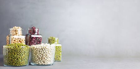 Bowls and jars of various legumes: chickpeas, lentils, mung, red and white bean on a gray background with copy space. Banner.の写真素材