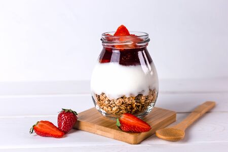 Jar of granola with yogurt, strawberry jam and fresh strawberries on a white wooden background. Healthy vegan breakfast concept.の写真素材