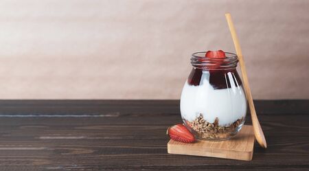Jar of granola with yogurt, strawberry jam and fresh strawberries on a brown wooden background. Healthy vegan breakfast concept. Banner with copy space.の写真素材