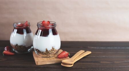Jars of granola with yogurt, strawberry jam and fresh strawberries on a brown wooden background. Healthy vegan breakfast concept. Vegetarian food. Banner with copy space.の写真素材