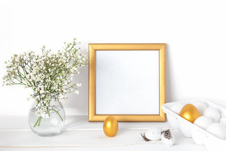 White and gold Easter eggs, a mockup with a gold frame and spring flowers in a vase on a white wooden table.の写真素材