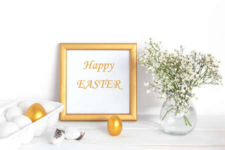 White and gold Easter eggs, a mockup with a gold frame and spring flowers in a vase on a white wooden tableの写真素材
