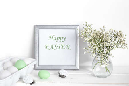 White and green Easter eggs, a mockup with a silver frame and spring flowers in a vase on a white wooden table.の写真素材