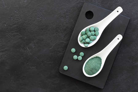 Green algae in powder and pills - chlorella, spirulina in white spoons on a black background. Healthy green food supplement concept.の写真素材