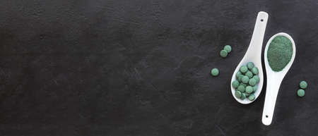 Green algae in powder and pills - chlorella, spirulina in white spoons on a black background. Healthy food concept.の写真素材